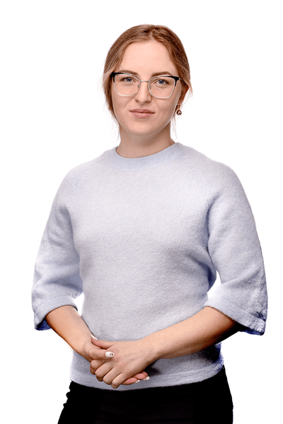 Portrait of a woman with light brown hair and glasses, wearing a pale blue sweater, standing with hands clasped in front.