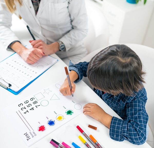 Young boy colors shapes on a worksheet with markers while a clinician in a white coat supervises at his desk.